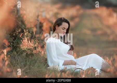 Giovane ragazza incinta in bianco abito lungo si siede su una radura verde. Foto Stock