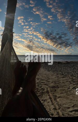 Di piedi di un turista in un'amaca sulla spiaggia, Xpuha Messico. Sullo sfondo ci sono il cielo nuvoloso all'alba, la spiaggia e il mare calmo. Concetto di viaggio A. Foto Stock