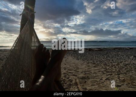 Primo piano dei piedi di un turista che si rilassa mentre si guarda al mare, in un'amaca su una spiaggia tropicale messicana all'alba. Sullo sfondo il cielo nuvoloso. Foto Stock