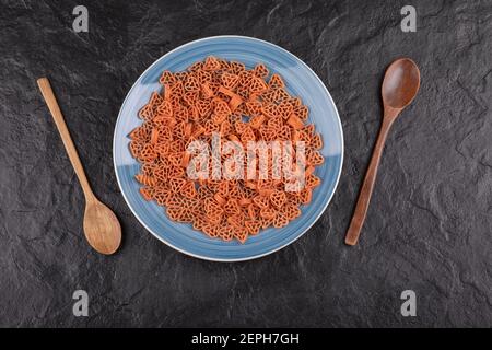 Un piatto di pasta cruda a forma di cuore con legno cucchiai su tavolo nero Foto Stock