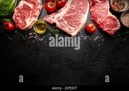 Varietà di bistecche fresche di carne cruda Black Angus prime T-Bone, New York, Ribeye e stagionatura su sfondo nero, vista dall'alto Foto Stock