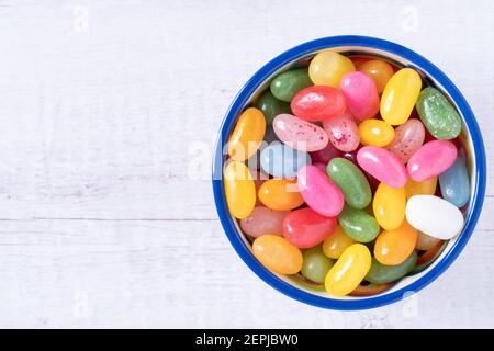 Fagioli di gelatina dolci in una ciotola di ceramica su uno sfondo di legno bianco con spazio di copia. Caramelle colorate con sapore di frutta mista. Vista dall'alto, primo piano. Spia An Foto Stock