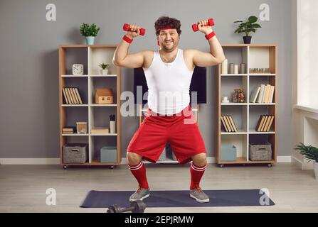 Divertente uomo chubby che si esercita con manubri in piedi su tappetino fitness in soggiorno Foto Stock