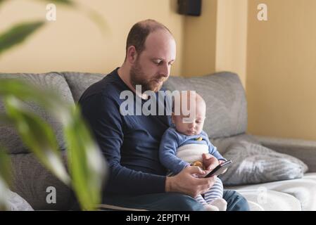Il padre caucasico che parla sul telefono mobile per lavorare in linea con il suo bambino neonato. Papà singolo che si prende cura di suo figlio. Famiglia felice e lavoro a casa Foto Stock