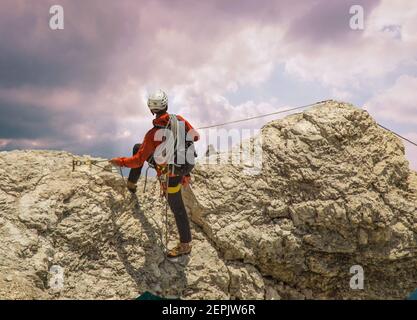 Un arrampicatore di roccia in una giacca rossa con le sue funi e l'attrezzatura da arrampicata segue la scena sottostante dalla cima della sua salita Foto Stock