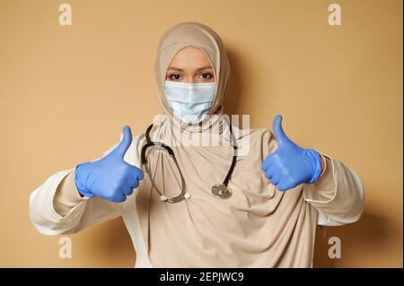 Medic giovane che indossa hijab e maschera medica mostrando pollici su gesturing con le mani in guanti medici di protezione. Sfondo beige con spazio per la copia Foto Stock