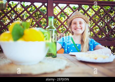 bambino moderno sorridente in tuta blu e cappello con verde bottiglia di limonata e piatto di limoni dell'azienda agricola locale seduto al tavolo con pranzo nel ter Foto Stock