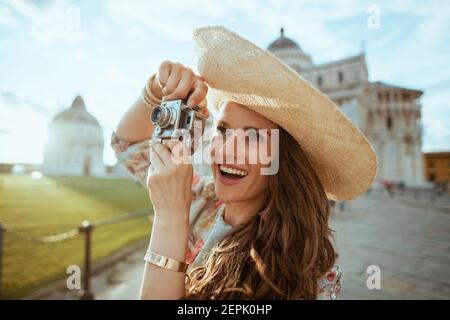 Sorridente elegante donna turistica solista in abito floreale con fotocamera retrò e cappello vicino alla Cattedrale di Pisa. Foto Stock