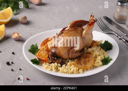 Quaglie cotte al forno con limone e arancia servite con il bulgur su un piatto bianco su sfondo grigio chiaro. Foto Stock