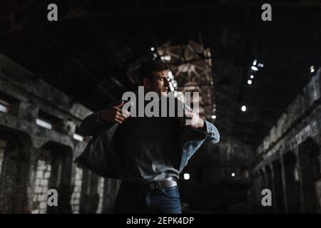 Uomo brutale ed elegante con giacca in jeans in piedi al buio vecchio edificio e che si affaccia sul lato Foto Stock