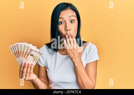 Bella donna ispanica che tiene 5000 sud coreana ha vinto banconote coprendo bocca con la mano, scioccata e impaurita per errore. Sorpresa espressione Foto Stock