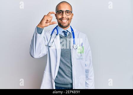 Uomo ispanico adulto che indossa uniforme medico e stetoscopio sorridente e. gesturing sicuro con la mano che fa piccolo segno di taglia con le dita guardare e il Foto Stock