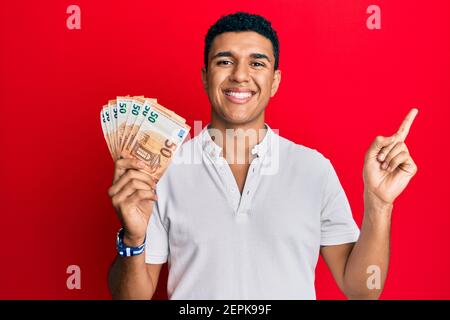 Giovane arabo che detiene 50 banconote in euro sorridendo felice di indicare con la mano e il dito di lato Foto Stock