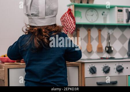 Bambina caucasica, indossando un cappello da chef, giocando ad essere una cuoca nella sua piccola cucina giocattolo. Foto Stock