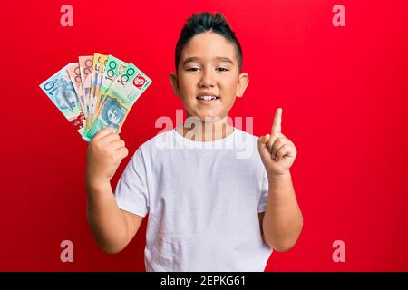 Piccolo ragazzo ispanico ragazzo che tiene dollari australiani sorridendo con un'idea o una domanda che punta il dito con il volto felice, numero uno Foto Stock