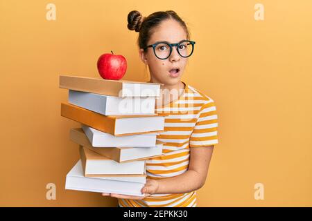 Bella bruna ragazza piccola che tiene un mucchio di libri con una mela in cima in faccia d'urto, guardando scettico e sarcastico, sorpreso con il mo aperto Foto Stock