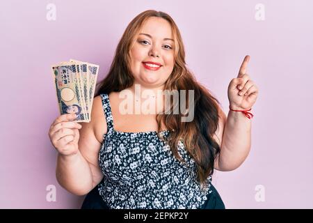 Più taglia giovane donna caucasica che tiene le banconote giapponesi yen sorridenti felice di puntare con mano e dito a lato Foto Stock