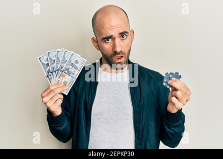 Giovane ispanico che gioca a poker tenendo banconote e chip dollari in faccia shock, guardando scettico e sarcastico, sorpreso con bocca aperta Foto Stock