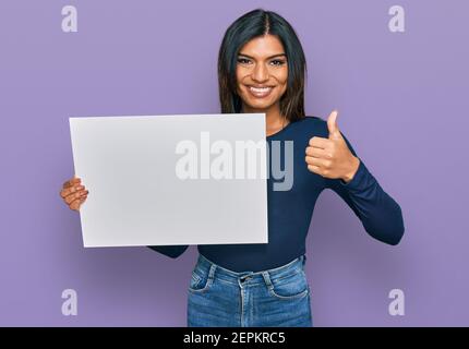 Giovane donna transessuale latina che tiene vuoto banner vuoto sorridendo felice e positivo, pollice su facendo eccellente e segno di approvazione Foto Stock