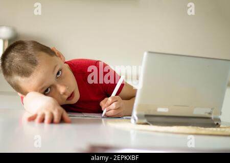 Il ragazzo stanco 6-10 completa le lezioni scolastiche e i compiti online in cucina, guardando il computer tablet. Il concetto di istruzione a distanza. Soggiorna in Foto Stock