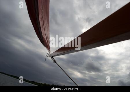 Primo piano del ligneo (braccio) su un piccolo gommone con una vela rossa e corde bianche. Con una silhouette che si staglia su un cielo grigio chiaro Foto Stock