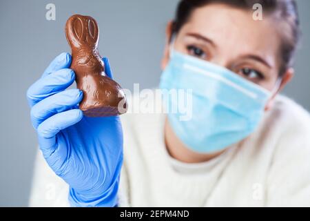 Triste giovane donna che indossa maschera protettiva e guanti medici che tengono un coniglietto di Pasqua cioccolato, espressione triste e depresso, vacanza in casa isolat Foto Stock