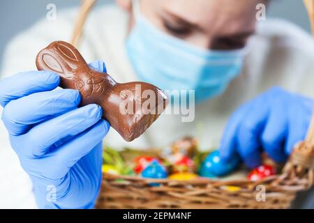 Persona che indossa blu protettivo guanti di gomma di lattice medico che tengono coniglietto di Pasqua di cioccolato, organizzando decorazione festiva e uova colorate in cestino, C. Foto Stock