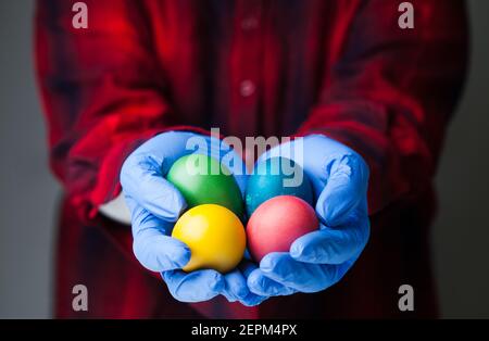 Persona che indossa guanti di protezione medica blu che tengono uova di Pasqua colorate, vacanza in mezzo globale Coronavirus COVID-19 malattia da virus pandemia, in tutto il mondo Foto Stock