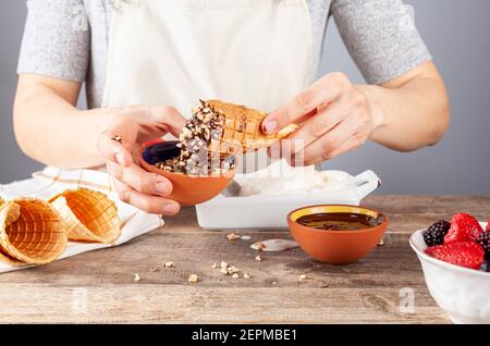 Una chef donna sta immergendo i coni fatti in casa per gelato al waffle in pezzi di cioccolato fuso e noce per un look artigianale. Altri coni laminati a mano sono lì. A m Foto Stock