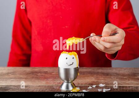 Un bambino piccolo sta mangiando l'uovo di humpty dumpty bollito morbido messo in tazza dell'uovo su un tavolo di legno della colazione. Lei usa il cucchiaio per mangiare l'uovo dal suo guscio. Lei Foto Stock