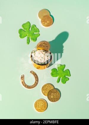 Concetto di giorno di San Patrizio. Vita moderna con simboli fortunati e cibo dolce per la festa di San Patrizio. Cuppake di velluto verde, shamrock, monete d'oro e ferro di cavallo su sfondo verde. Verticale Foto Stock