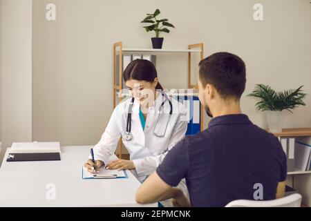 Amichevole praticante sorridendo e scrivendo la prescrizione per il giovane maschio paziente Foto Stock