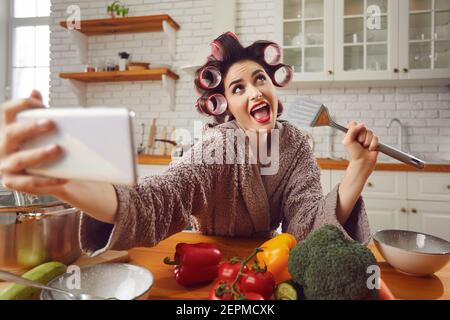 Donna in capelli arricciacapelli prendendo divertente selfie su cellulare mentre cucina in cucina Foto Stock