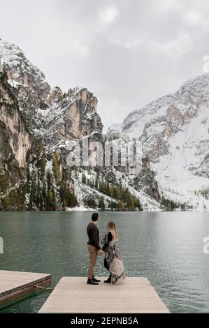 Corpo pieno di coppia dolce amorevole che si abbracciano l'un l'altro Molo in legno contro il lago di Braies circondato da neve montagne Foto Stock