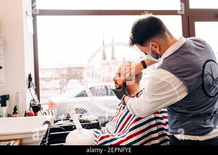 Vista posteriore di barbiere maschile anonimo su maschera facciale rasatura della barba dell'uomo utilizzando un rasoio dritto nel salone di bellezza durante la pandemia del coronavirus Foto Stock