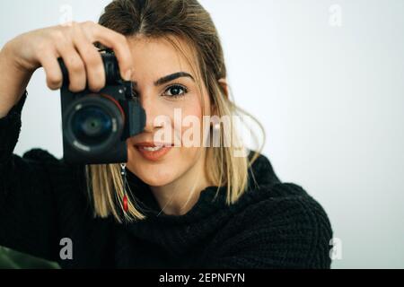 Crop femmina sorridente in maglione nero guardando la fotocamera mentre scattare foto con una fotocamera digitale professionale in casa su bianco sfondo Foto Stock