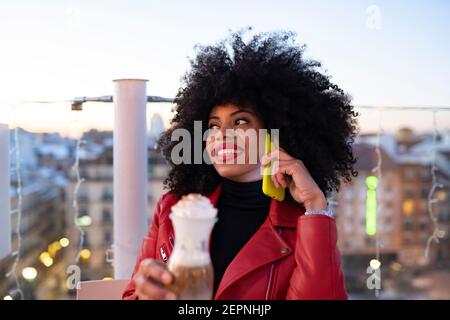 Allegra donna afroamericana con acconciatura Afro sorridente luminoso mentre seduto sul balcone con un bicchiere di bevanda e parlando telefono cellulare Foto Stock