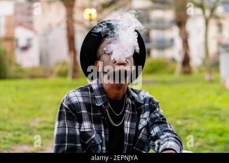 Serious African American maschio indossando occhiali da sole eleganti camicia a scacchi e. cappello fumando sigaretta Foto Stock