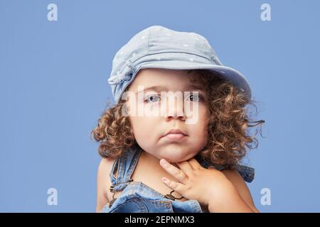 Adorabile bambino sconvolto con occhi marroni che indossano abiti casual e cappello su sfondo blu guardando via Foto Stock