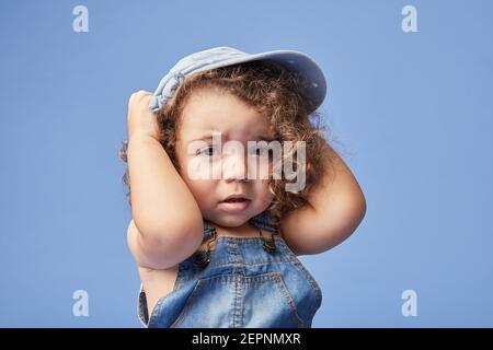 Adorabile bambino sconvolto con occhi marroni che indossano abiti casual e cappello su sfondo blu guardando la fotocamera Foto Stock