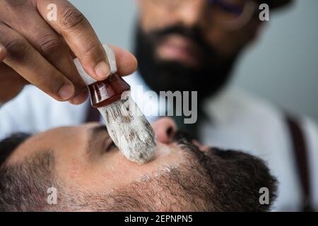 Primo piano di crop maschile etnico beauty master applicando una rasatura liscia sapone sulla faccia di uomo bearded usando la spazzola in acconciatura salone Foto Stock