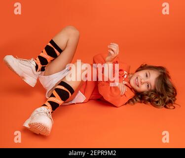 Bambina bella ragazza 6-7 anni bambino sdraiato sul pavimento in studio in fondo vestiti arancione. È felice, ridendo e gioisce. Infanzia concep Foto Stock