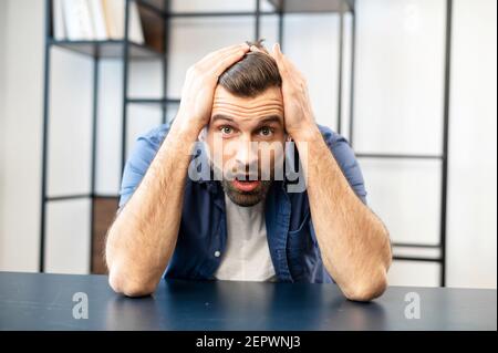 Un giovane dipendente di tipo con portatore di hipster travagliato con bocca aperta è seduto alla scrivania e tenendo la testa in mano, sentendosi frustrato e spaventato da problemi occured, scioccato per sentire le notizie difettose inattese Foto Stock
