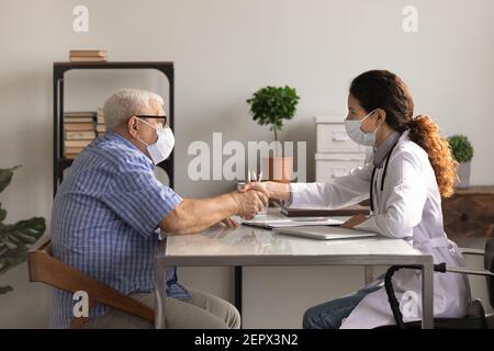 Vista laterale medico e paziente maturo che indossa maschere scuotendo le mani Foto Stock