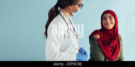 Donna musulmana felice guardando medico femminile e sorridendo su sfondo blu. Donna in clinica medica per la vaccinazione. Foto Stock