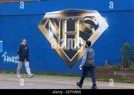 Londra, UK, 28 febbraio 2021: Un murale a Tulse Hill raffigura il logo NHS nello stile di un badge supereroe. Passers-by prendere l'esercizio o la coda per il negozio Co-op dietro l'angolo, alcuni indossando maschere viso e tutti socialmente distanziando. Anna Watson/Alamy Live News Foto Stock