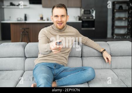 Eccitato giovane uomo hipster bearded con bocca aperta seduta sul divano nel soggiorno, puntando un telecomando tv sulla telecamera, cambiando canali, felice con un programma televisivo, guardando film Foto Stock