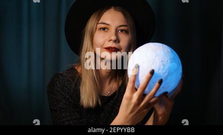 Una giovane donna che teller fortuna in un cappello sta tenendo una palla magica. Bella donna psichica sta indovinando con una sfera magica blu Foto Stock