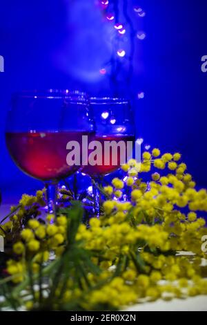 Mimosas e due bicchieri di vino rosso su un blu sfondo con luci offuscate a forma di cuori piccoli Foto Stock