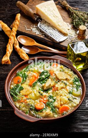 zuppa di kale di pollo con anellini di pasta e verdure in una ciotola di argilla su tavola di legno scuro con grissini e parmigiano, vista verticale, c Foto Stock